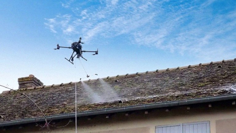 Drone AltaClean DJI Matrice 350 RTK en pulverisation active du biocide AMM, gros plan sur le drone et le jet de pulverisation au-dessus de la toiture moussee